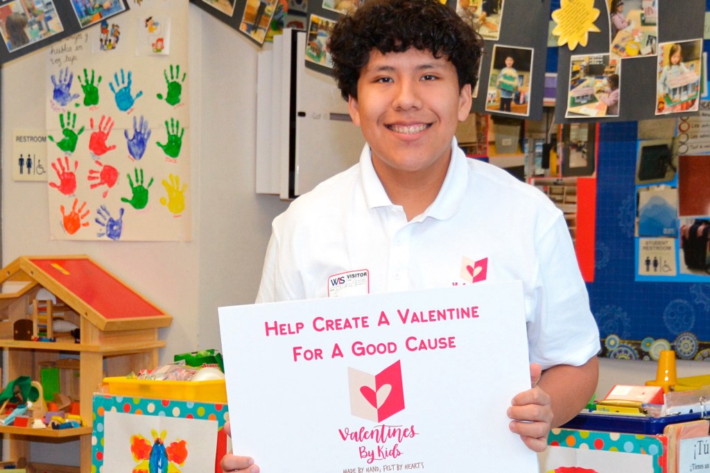 A smiling teenage boy holds a sign that says “Help create a valentine for a good cause - Valentines By Kids”