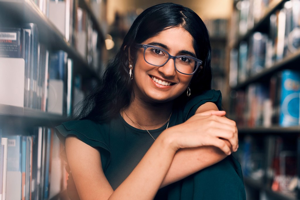 A smiling teenage girl wearing glasses sitting in a library.