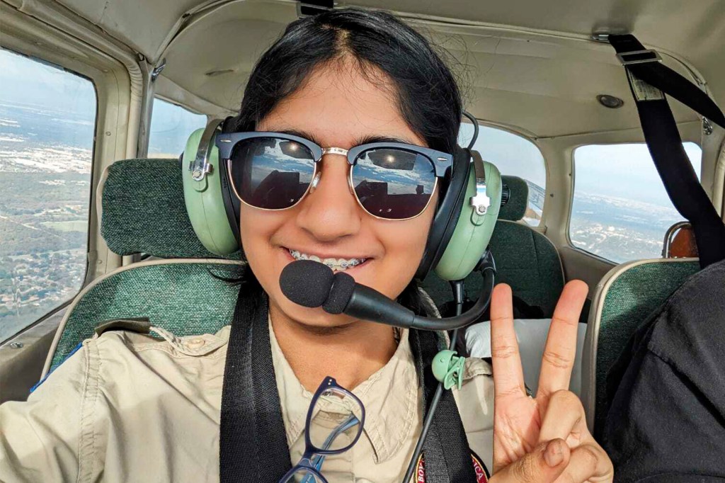 A smiling teenage girl wearing sunglasses in the cockpit of a plane, holding up the peace sign.
