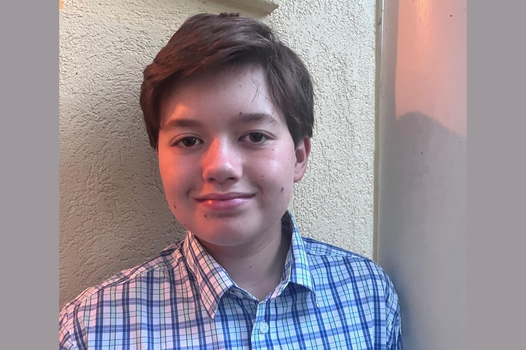 Portrait of a smiling boy wearing a blue and white plaid shirt, standing against a light-colored wall.
