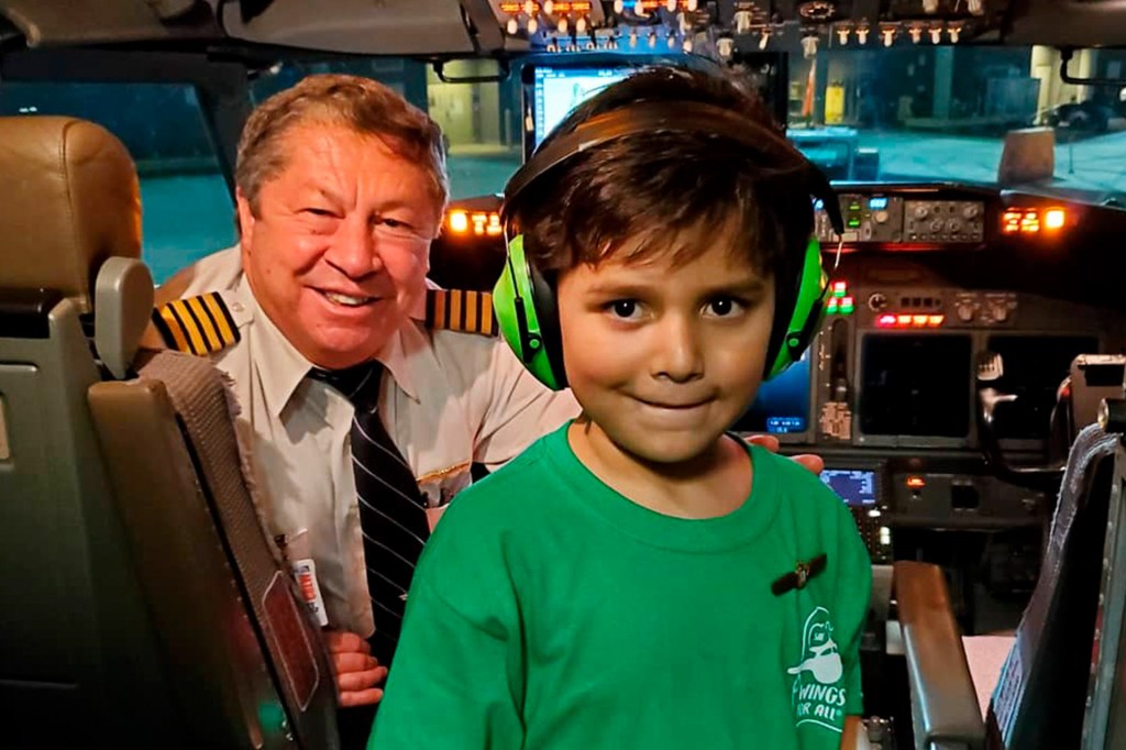 A boy wearing green headphones and a green t-shirt standing in the cockpit of a plane. The pilot sits behind him.