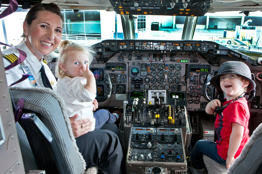 A young boy sits in a pilot seat of a parked plane. A member of the flight crew holds a toddler on her lap.