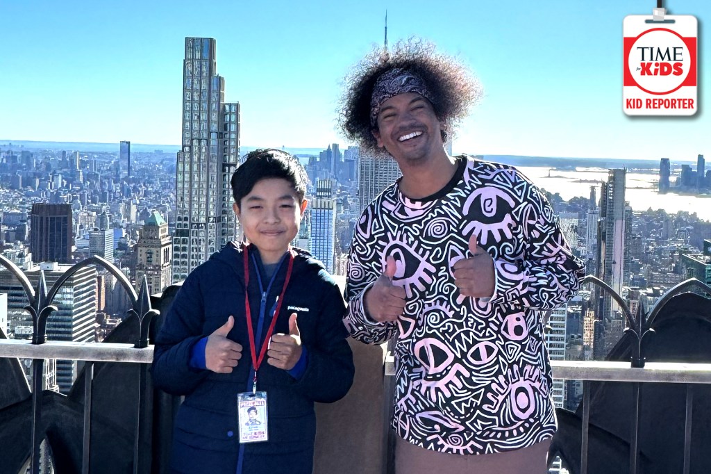 A smiling man and boy stand outdoors on a tall observation deck overlooking New York City. Both give thumbs-up gestures, and the boy wears a TIME for Kids Kid Reporter badge.