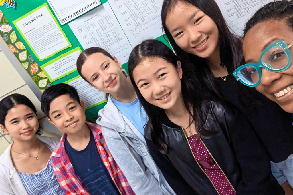 A group of students and a teacher smile for a photo in a classroom, standing in front of a green bulletin board with project papers displayed.