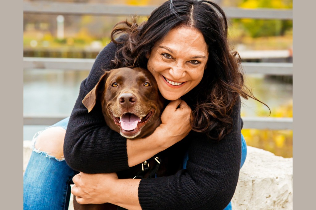 Smiling woman hugs a brown dog outdoors, both looking at the camera.