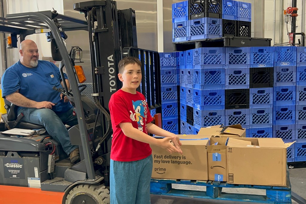 Smiling boy in a red shirt stands beside a man driving a forklift, with boxes stacked on a blue pallet and tall stacks of blue crates behind them.