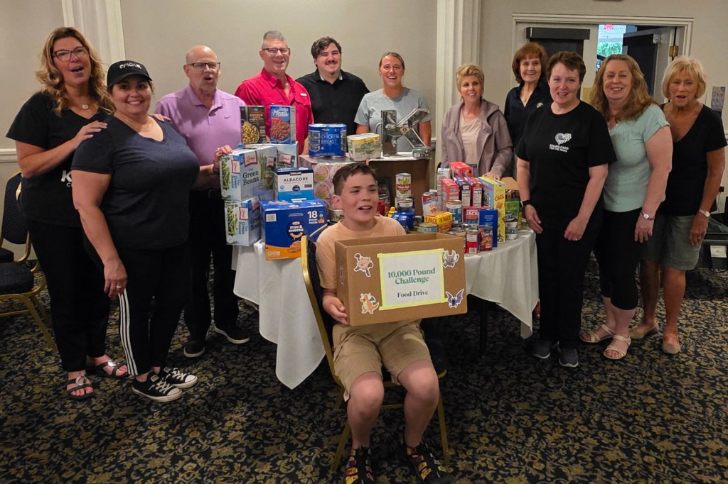 Smiling boy sits holding a box labeled “10,000 Pound Challenge Food Drive,” surrounded by adults standing near a table filled with canned goods and food donations.