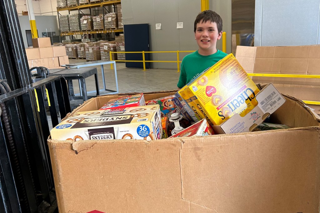 Smiling boy in a green shirt stands next to a large box filled with food items in a warehouse.