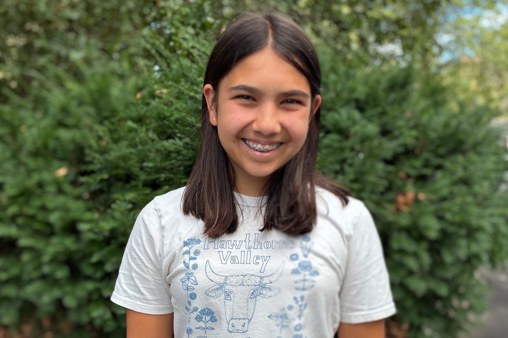 ortrait of a smiling girl with straight dark hair wearing a white “Hawthorne Valley Farm Camp” T-shirt, standing outdoors in front of green bushes.