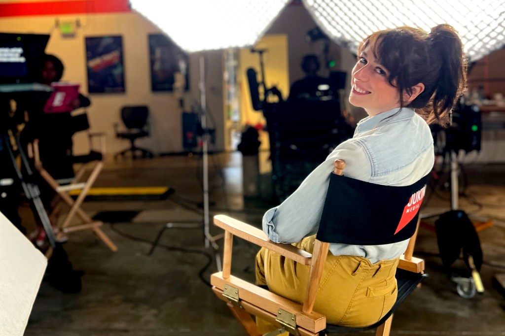 A person smiles at the camera while sitting in a director’s chair on a film set. The chair has a red and white logo on the back, and bright studio lights and camera equipment are visible in the background.