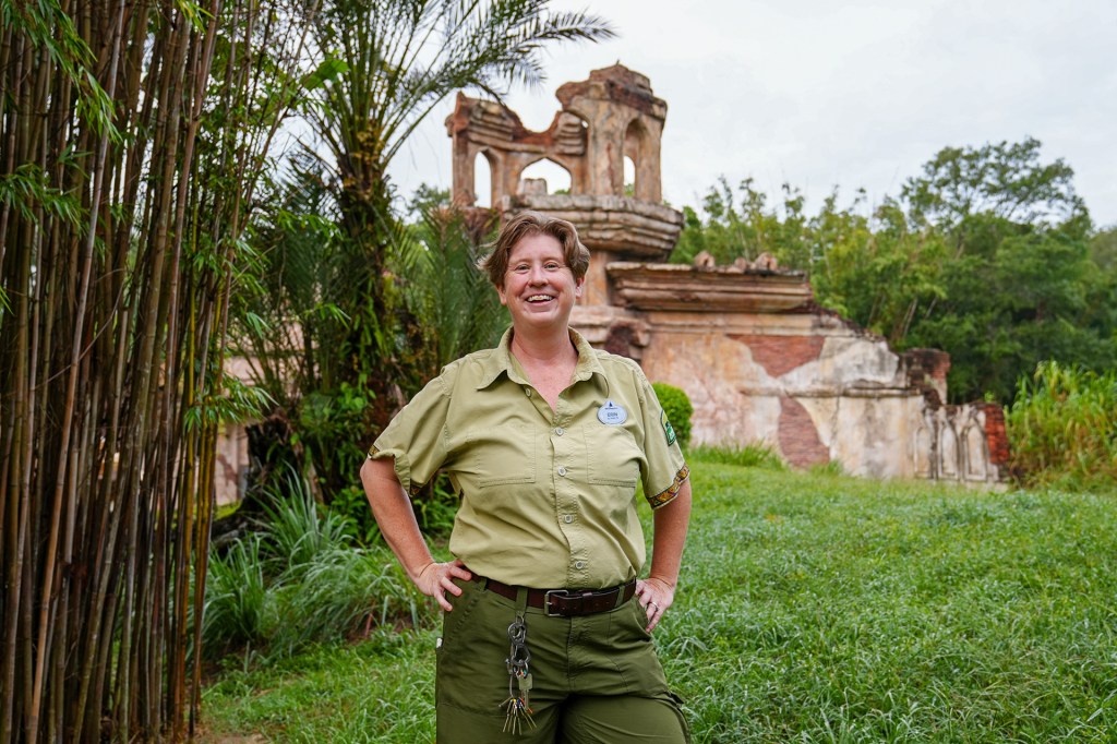 A smiling wildlife worker stands outdoors in uniform, surrounded by greenery and old stone ruins.