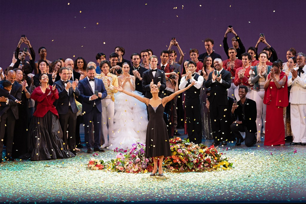 Ballerina stands on stage surrounded by confetti and flowers as a crowd of performers and audience members applaud behind her.