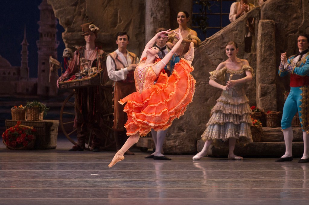 A ballerina in an orange dress leaps gracefully on stage during a performance, with dancers in period costumes watching in the background.