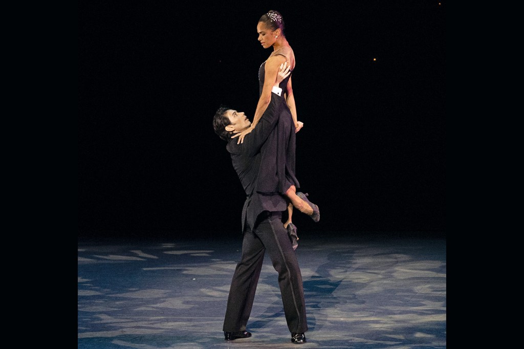 Ballet dancer in a black dress is lifted into the air by a male dancer during a stage performance.