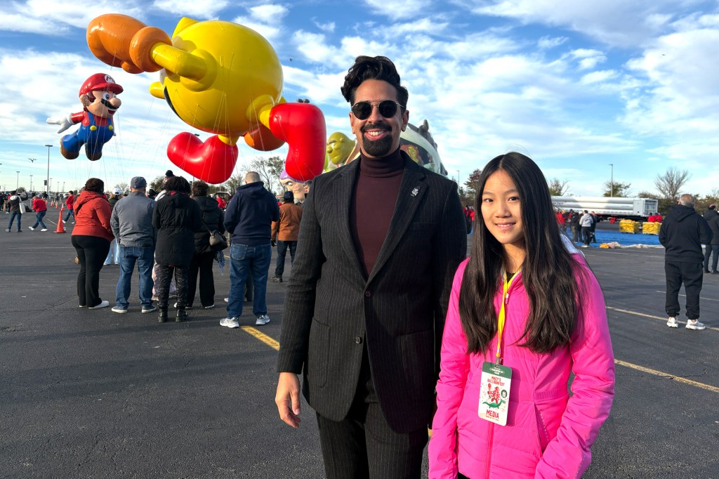 A smiling man in a dark suit and sunglasses stands beside a girl in a bright pink jacket at a parade balloon setup area, with giant Mario and Pac-Man balloons behind them.